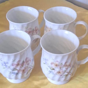May include: A set of four white ceramic mugs featuring a subtle vertical ribbed texture and a delicate floral pattern in soft shades of pink, blue, and green. Each mug has a rounded body, a flared rim, and a curved handle on the side.