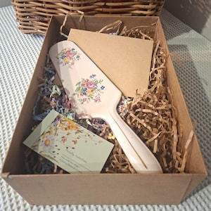 May include: A white ceramic cake server with a floral pattern featuring pink, yellow, and blue flowers, nestled in a brown cardboard gift box filled with shredded paper, accompanied by a blank tan card and a small rectangular business card.