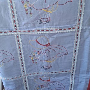 May include: A light blue quilt featuring repeating embroidered panels of a figure wearing a wide-brimmed hat and holding a bouquet of colorful flowers. The panels are separated by white lace trim woven with thin red ribbon accents throughout.