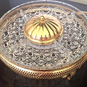 May include: A round clear glass divided serving dish with a starburst pattern, featuring a central gold-toned lidded bowl. The glass dish is set within a decorative gold-toned metal frame with two side handles and an intricate lattice border.