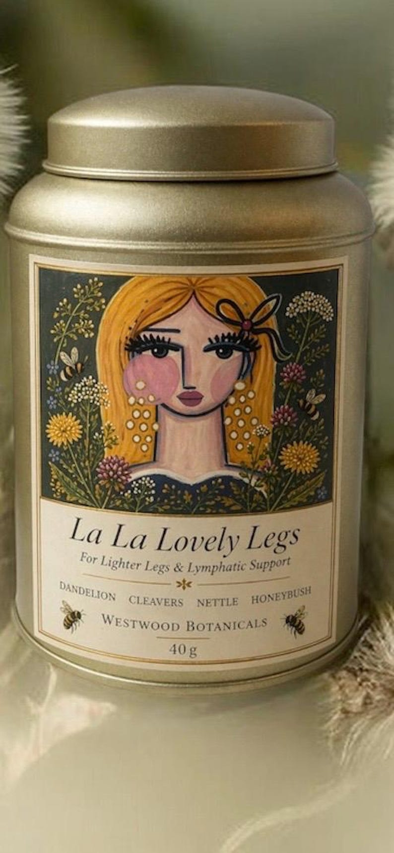 May include: A gold-colored metal tin with a decorative label featuring a stylized illustration of a woman with blonde hair and floral accents. The label reads "La La Lovely Legs" and lists botanical ingredients. The tin contains 40g of product.