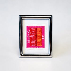 Könnte beinhalten: Ein rechteckiger, silberfarbener Metallrahmen mit einer Briefmarke. Die Briefmarke ist leuchtend pink mit orangefarbenen und weißen grafischen Elementen, dem Text "DEUTSCHE BUNDESPOST", "40", "HEINRICH SCHÜTZ + 6.11.1672" und einer weißen Schreibschrift-Signatur.