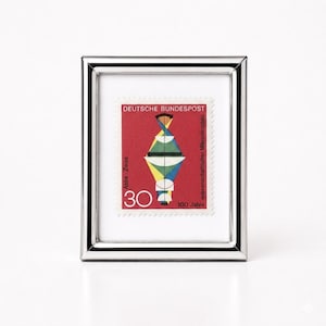 May include: A rectangular silver-toned metal frame containing a vintage postage stamp. The stamp features a red background with a geometric abstract design, the text DEUTSCHE BUNDESPOST, Abbe-Zeiss, 30, and wissenschaftlicher Mikroskopbau 100 Jahre.