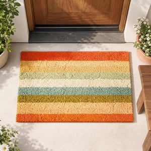 Pode incluir: Um tapete retangular com listras horizontais em tons de laranja, amarelo, verde e azul. O tapete é feito de um material texturizado e é colocado em frente a uma porta de madeira.