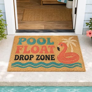 以下が含まれることがあります： 「POOL FLOAT DROP ZONE」の文字がターコイズ、オレンジ、黒で書かれた長方形のドアマット。右側にはフラミンゴとヤシの木があります。マットは天然繊維素材で作られています。