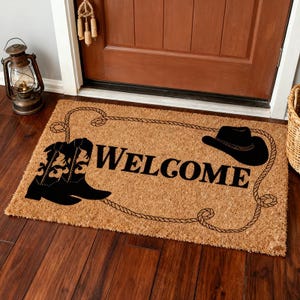 Może przedstawiać: Prostokątna wycieraczka w stylu kowbojskim. Mata jest beżowa z czarną obwódką ze sznura i napisem "WELCOME" dużymi czarnymi literami. Na macie znajduje się czarny kowbojski but i kapelusz.