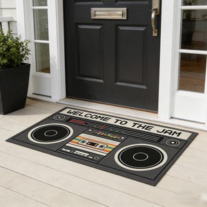 Op de afbeelding: Een rechthoekige deurmat met een retro boombox ontwerp. De mat is voornamelijk grijs met zwarte en witte accenten, met een cassettebandje en de woorden "WELCOME TO THE JAM".