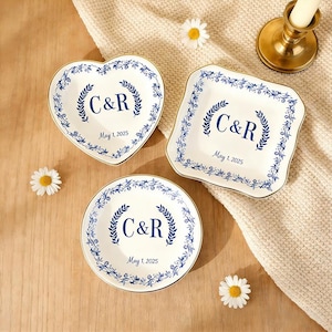 May include: Three white ceramic dishes with blue floral designs and gold rims. Each dish has the initials "C&R" and the date "May 1, 2025" in blue. The dishes are in heart, square, and round shapes.