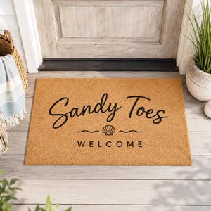 Könnte beinhalten: Eine rechteckige Fußmatte aus natürlicher Kokosfaser. Auf der Matte steht in Schreibschrift "Sandy Toes", mit einer Muschelgrafik und dem Wort "Welcome" darunter. Die Matte liegt vor einer Holztür.