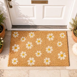 May include: A rectangular doormat with a tan background and a pattern of white daisies with yellow centers. The mat is placed in front of a white door with a brass mail slot. Potted plants are on either side.