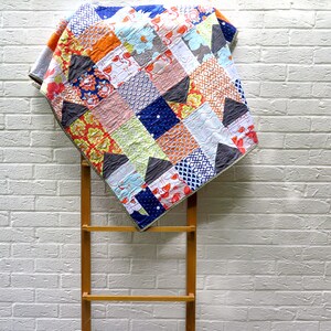 May include: A colourful patchwork quilt with a variety of patterns and colours, including orange, blue, green, and white. The quilt is draped over a wooden ladder against a white brick wall.