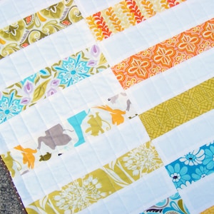 May include: A white quilt with colourful fabric strips sewn together in a horizontal pattern. The fabric strips feature various floral and geometric designs in shades of blue, green, orange, and yellow.