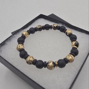 May include: A black beaded bracelet with gold-colored accents. The bracelet is made of round, porous black beads interspersed with gold-colored, textured beads. The bracelet is presented in a black gift box with a white interior.