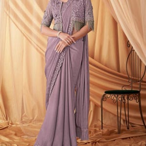 May include: A lavender saree with detailed embroidery and a matching bolero jacket. The jacket has delicate beadwork and fringe trim. The ensemble is draped elegantly, showcasing the flowing fabric and design.