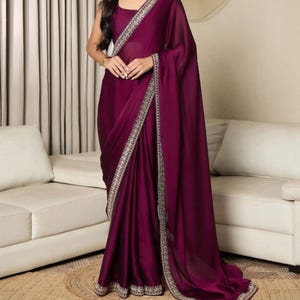 Può includere: Un sari viola scuro con una blusa abbinata. Il sari presenta un bordo decorativo argentato e dorato. Il tessuto drappeggia elegantemente, creando morbide pieghe. L'outfit è adatto per eventi formali.