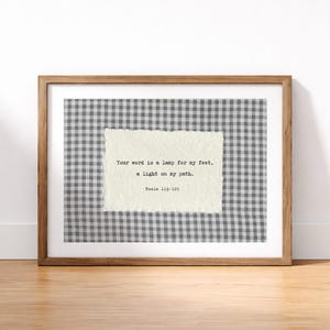Op de afbeelding: Houten ingelijst kunstwerk met een citaat uit Psalm 119:105. De tekst, "Uw woord is een lamp voor mijn voet, een licht op mijn pad", staat op getextureerd papier, tegen een grijs en wit geruit patroon. De lijst is van hout.