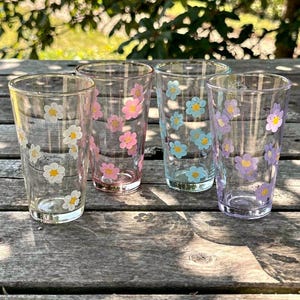 Puede incluir: Cuatro vasos transparentes decorados con patrones de margaritas coloridas. Cada vaso presenta un color diferente de margarita: blanco, rosa, azul y morado. Los vasos están colocados sobre una superficie de madera, con un fondo verde borroso.
