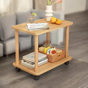 May include: A light wood-tone rolling side table with two shelves. The top shelf holds a glass of water with a lime, a magazine, a vase with flowers, and oranges. The bottom shelf has books and a wicker basket.