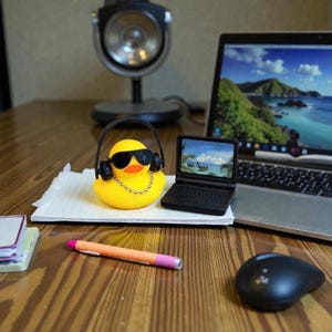 May include: A yellow rubber duck wearing black sunglasses, a silver ball chain necklace, and black headphones. Next to it is a miniature black laptop, a stack of white paper, an orange pen, a black computer mouse, and a desk lamp.