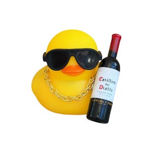 May include: A yellow rubber duck wearing black sunglasses and a gold chain necklace, holding a bottle of red wine. The wine bottle has a white label with red text that reads "Casillero del Diablo".