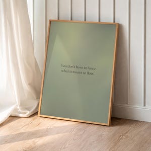 Könnte beinhalten: Ein gerahmter Kunstdruck mit einem salbeigrünen Hintergrund und einem Holzrahmen. Der Druck zeigt den Text: "You don't have to force what is meant to flow." Das Kunstwerk lehnt an einer weißen Wand und einem weißen Vorhang.
