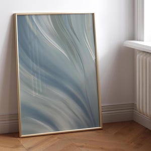 Op de afbeelding: Abstracte kunstafdruk in een gouden lijst. Het kunstwerk toont vloeiende, gelaagde penseelstreken in tinten blauw, grijs en wit. De ingelijste kunst leunt tegen een witte muur op een houten vloer.