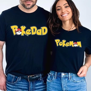 Puede incluir: Dos camisetas negras con el texto "PokeDad" y "PokeMom" en amarillo y rojo, con un diseño de estilo caricatura. Las camisetas son usadas por dos personas, combinadas con vaqueros azules.