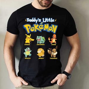 May include: Black t-shirt featuring the text "Daddy's Little Pokémon" above cartoon images of Pikachu, Bulbasaur, Charmander, Squirtle, Psyduck, and Eevee, each with a name below. The shirt is a short-sleeved crew neck.