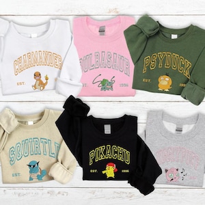 May include: A collection of six sweatshirts in various colors, each featuring a different Pokémon character. The sweatshirts display the character's name in a collegiate-style font, along with an image of the character. The sweatshirts are white, pink, green, tan, black, and gray.