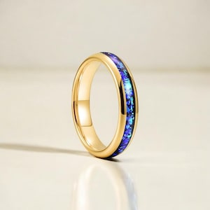 Op de afbeelding: Een goudkleurige ring met een band van blauw en paars iriserend materiaal. De ring staat tegen een neutrale achtergrond, waardoor de levendige kleuren van de inleg worden benadrukt. De ring is een sieraad.