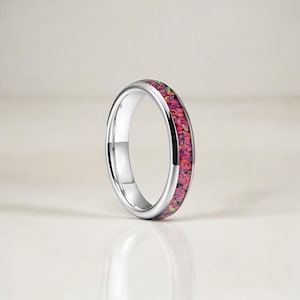 Op de afbeelding: Een zilveren ring met een band van levendig roze en rood ingelegd materiaal. De ring heeft een gepolijst, reflecterend oppervlak en het ingelegde materiaal lijkt te bestaan uit kleine, kleurrijke fragmenten.