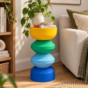 May include: A cylindrical side table composed of four stacked, rounded geometric segments in yellow, green, light blue, and dark blue. A white ceramic vase with a green plant and a book titled The Kinfolk Home are placed on the top surface.