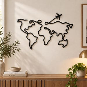 May include: A black metal wall art piece depicting a continuous line drawing of a world map. The design features simplified continental outlines and a small airplane silhouette trailing a line above the landmasses, mounted on a plain wall.