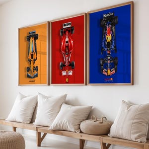 May include: Three framed Formula 1 car prints. The prints feature overhead views of race cars against orange, red, and blue backgrounds. The frames are wood-toned, and the prints are displayed above a wooden bench with white pillows.
