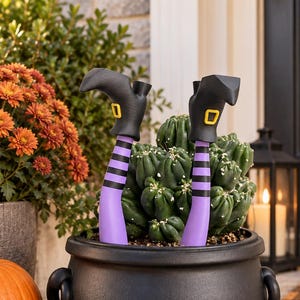 May include: A black cauldron planter containing a green cactus and two decorative witch legs. The legs feature purple and black striped stockings, black boots with yellow buckles, and are positioned upright as if emerging from the plant.
