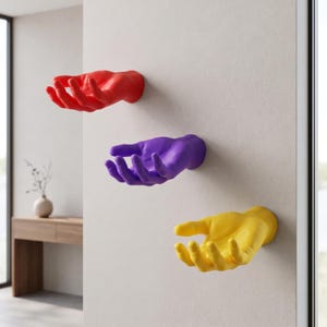 May include: Three wall-mounted hand sculptures arranged vertically. The top hand is bright red, the middle hand is vibrant purple, and the bottom hand is sunny yellow. Each hand is sculpted in an open, cupped position, extending outward from the wall.