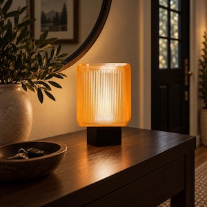 May include: A table lamp featuring a rectangular, amber-colored, ribbed glass shade that emits a warm glow. The shade is supported by a solid, square, black base. The lamp provides soft illumination, highlighting the vertical texture of the glass.
