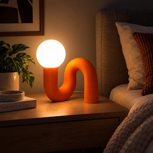 May include: A table lamp featuring a bright orange, curved, tubular base that forms an S-shape. A spherical, frosted white glass globe sits atop one end of the orange base, emitting a warm, soft glow that illuminates the surrounding area.
