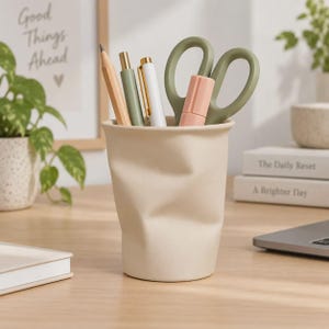 May include: A beige, crumpled-style desk organizer cup holding a wooden pencil, a sage green pen, a white pen with gold accents, a small pink marker, and sage green scissors. The cup has a matte finish and a soft, irregular, folded texture.