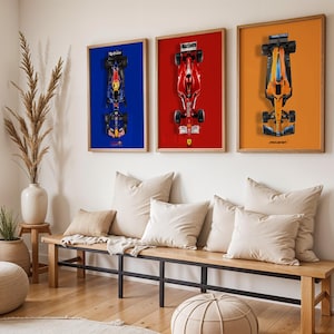 May include: Three framed Formula 1 car prints. The first features a blue car on a blue background, the second a red car on a red background, and the third an orange car on an orange background. The frames are wood-toned, and the prints are displayed above a wooden bench.