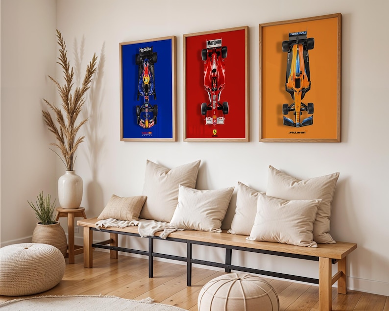 May include: Three framed Formula 1 car prints. The first features a blue car on a blue background, the second a red car on a red background, and the third an orange car on an orange background. The frames are wood-toned, and the prints are displayed above a wooden bench.