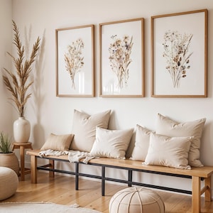 May include: Three framed botanical prints with dried flowers hang above a wooden bench with beige pillows. A large vase with tall dried grass and a woven ottoman complete the neutral-toned, natural decor.