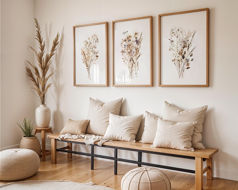 May include: Three framed botanical prints with dried flowers hang above a wooden bench with beige pillows. A large vase with tall dried grass and a woven ottoman complete the neutral-toned, natural decor.