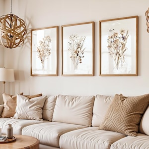 May include: Three framed botanical prints with dried flowers in a living room setting. The prints are in light brown wooden frames, and the couch has beige cushions and pillows. A wooden coffee table and decorative light fixtures complete the scene.