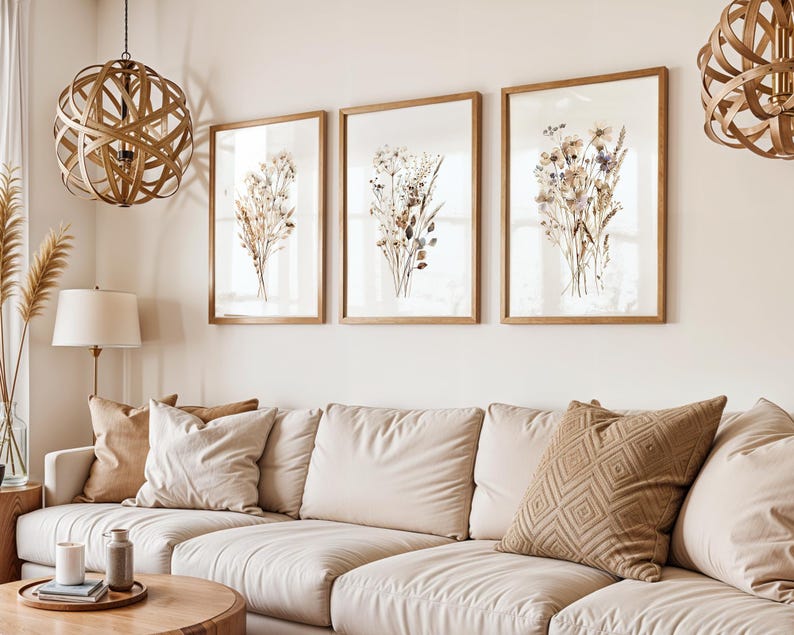 May include: Three framed botanical prints with dried flowers in a living room setting. The prints are in light brown wooden frames, and the couch has beige cushions and pillows. A wooden coffee table and decorative light fixtures complete the scene.