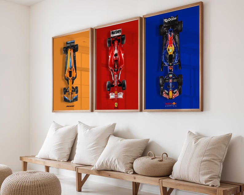 May include: Three framed Formula 1 car prints. The prints feature overhead views of race cars against orange, red, and blue backgrounds. The frames are wood-toned, and the prints are displayed above a wooden bench with white pillows.