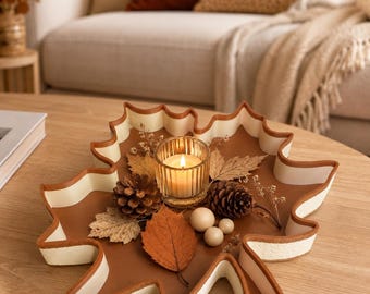May include: A maple leaf-shaped tray with a brown interior and white edges, containing a lit glass candle holder, two pinecones, three small wooden spheres, and several dried leaves, including one prominent orange leaf and gold-toned foliage.
