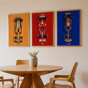 May include: Three framed Formula 1 race car prints. The prints feature overhead views of the cars against orange, red, and blue backgrounds. The frames are a light wood color. A round wooden table and chairs are in the foreground.