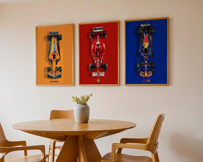 May include: Three framed Formula 1 race car prints. The prints feature overhead views of the cars against orange, red, and blue backgrounds. The frames are a light wood color. A round wooden table and chairs are in the foreground.