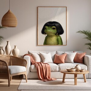 May include: A framed art print featuring a green frog with black hair and gold hoop earrings. The frog is the focal point, set against a neutral background. The artwork hangs above a white sofa with peach-colored pillows and a matching throw.